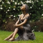 Bronzefigur einer sitzenden Frau auf einem Rasen im Garten